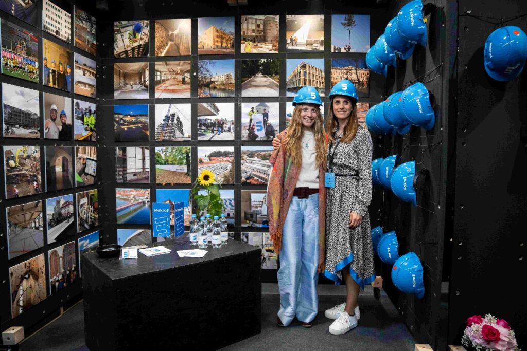 dvije djevojke s plavim kacigama na glavi stoje u prostoriji u kojoj na zidu visi mnogo plavih kaciga i fotografija